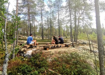 Tre personer sitter vid en rastplats på träbänkar.