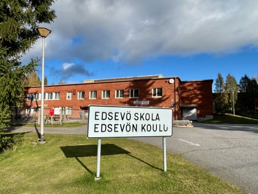 Kuvan taustalla Edsevön koulu. Se on punatiilinen rakennus. Etualalla on kyltti, jossa lukee "Edsevö skola, Edsevön koulu".