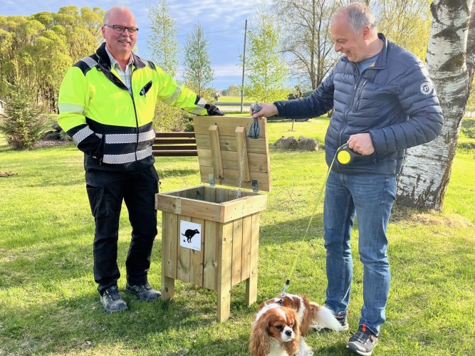 Kunnanpuutarhuri pitää koirankakka-astian kantta auki Ankkalammen kohdassa Pännäissä. Kunnan teknisten palveluiden päällikkö laittaa kakkapussin astiaan. Milou-koira seisoo nurmikolla koirankakkalaatikon edessä.