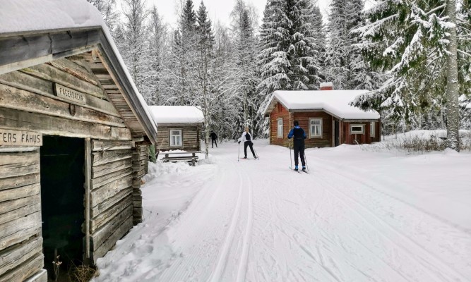 Fagerbacka skidspår. Tre skidåkare i spåret vid Fagerbacka fäbodställe. Två stugor och en lada syns på bilden, skog i bakgrunden. 