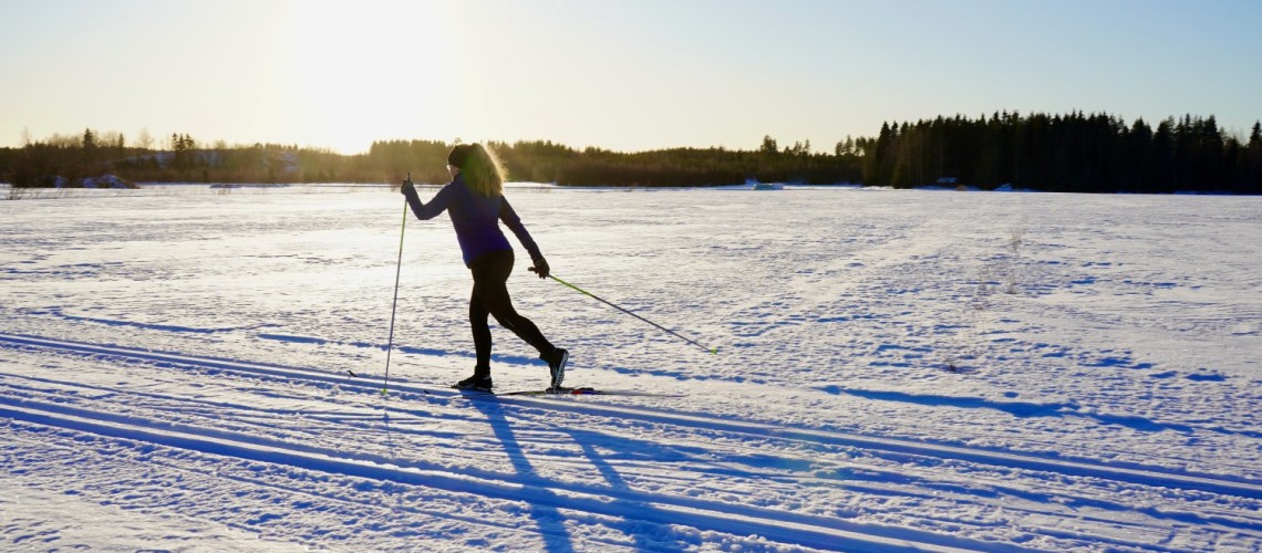 Yksinäinen hiihtäjä hiihtää latua pitkin kohti aurinkoa. Tunnelmallinen kuva.