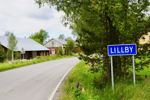 En landsväg går genom en gårdsgrupp. Till höger i bild finns en skylt med texten LILLBY. Det är sommar.