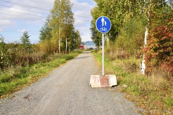 En grusväg med ett trafikmärke som berättar att det är en gång- och cykelväg. I bakgrunden kan man ana Amada Automations fabrik.