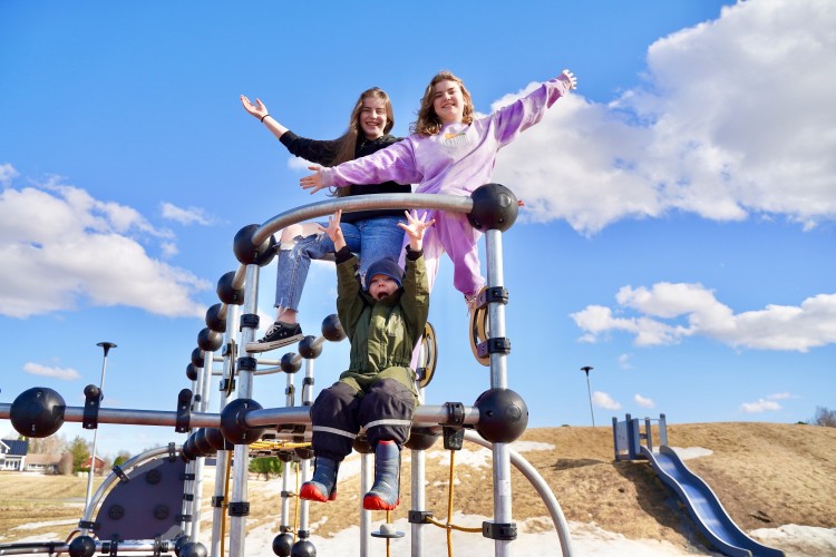 Två flickor står står i en klätterställning och ett barn sitter. Alla tre jublar med händerna mot skyn.
