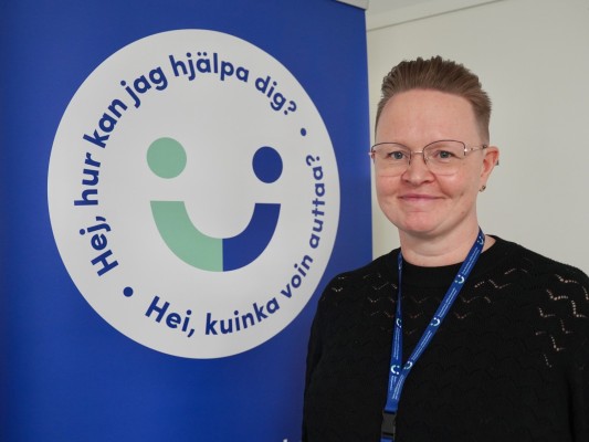 Seniorrådgivaren Camilla Passell står bredvid en skylt med Österbottens välfärdsområdes logo. I logon står det: Hej, hur kan jag hjälpa dig. Hei, kuinka voin auttaa.