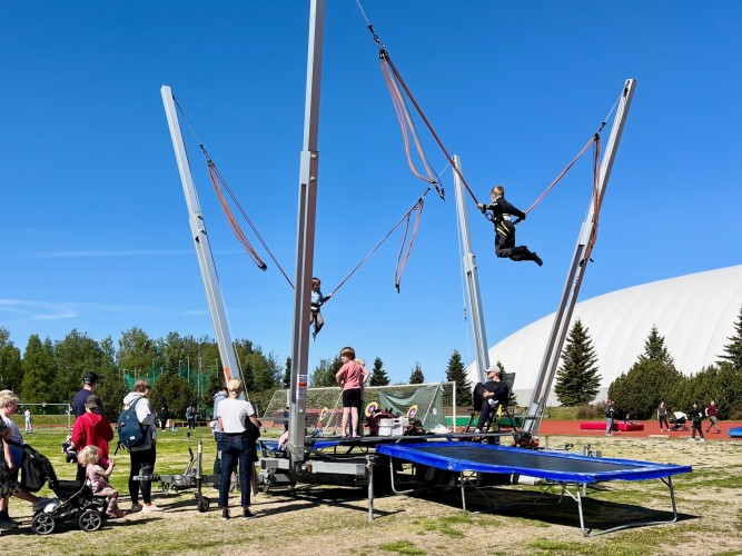 Ett barn hoppar i trampolinen, fastspänd i en lina. Folk köar bredvid.