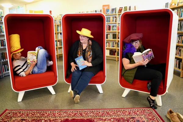 Tre kvinnor sitter i läsfåtöljer i biblioteket. Alla tre läser en bok medan de har en rolig hatt på huvudet.