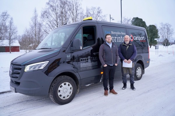 Två manliga taxichaufförer står utanför en taxibil och ler mot kameran. Taxidörren är öppen.