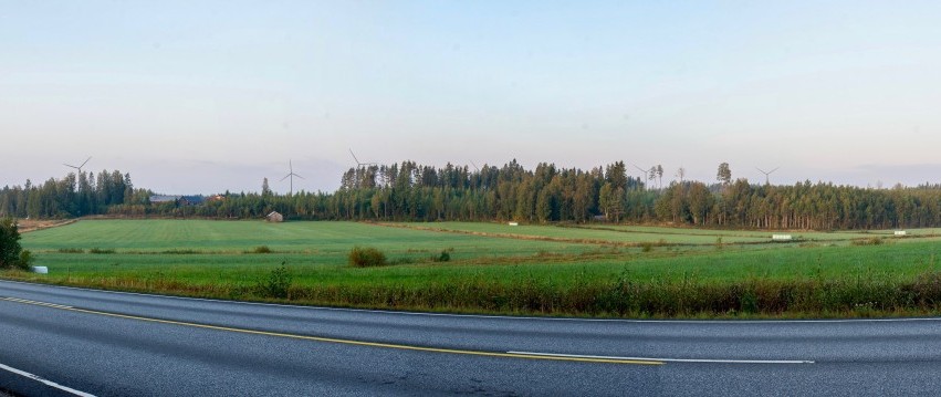 Ett fotomontage från Lappfors, där det syns vindkraftverk långt borta mellan träden. I förgrunden landsvägen och vyerna när man kör in i Lappfors från Essehållet.