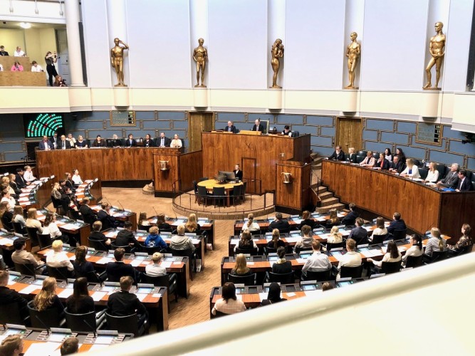 Bild inifrån riksdagshusets plenisal. Ungdomar sitter på riksdagesmännens platser. Talmannen är på plats längst fram i salen.