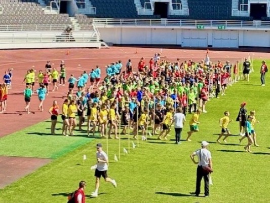 Massor av barn i färgglada kläder på gräsmattan i Olympiastadion. Solen skiner.